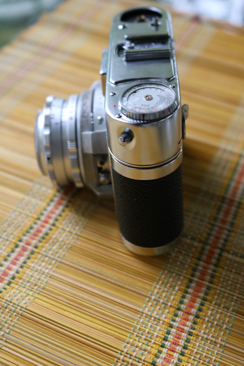 Pu&ograve; includere: Una macchina fotografica a pellicola da 35 mm argentata e nera con una impugnatura in pelle. La macchina fotografica ha un esposimetro incorporato e un obiettivo a messa a fuoco manuale.