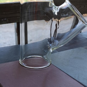 May include: A clear glass bong with a cylindrical body and an angled mouthpiece. The bong sits on a dark surface, with a blurred outdoor background. The glass is transparent, allowing a view through the piece.