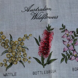 May include: A white linen tea towel featuring the text Australian Wildflowers in black script. The fabric displays botanical illustrations of yellow wattle, a red bottlebrush, and pink boronia flowers, each labeled with its name in black capital letters.