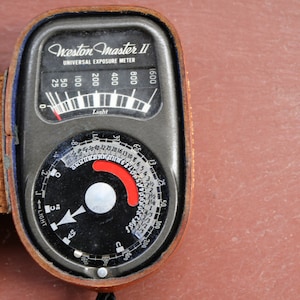 May include: Vintage Weston Master II Universal Exposure Meter in a leather case. The meter has a black face with white markings and a needle indicator. The case is brown leather with a blue interior. The meter reads "Light" and has a dial.