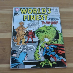 Può includere: Una copertina di fumetto con Superman e Batman che combattono una creatura aliena verde. Il testo della copertina recita "World's Finest Comics" e "The Sorcerer from the Stars!"