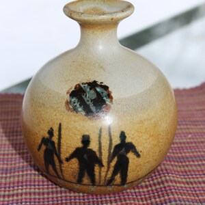 Peut inclure: Un vase en céramique brun avec un corps rond et un col étroit. Le vase est décoré d'un motif peint de trois personnages et d'un cercle avec un motif bleu et noir.
