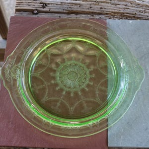 May include: A vintage, light green glass plate with a decorative floral pattern in the center and around the rim. The plate has two handles and is set on a brown and gray surface. The plate is likely from the Depression era.