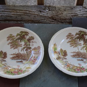 May include: Two antique, off-white ceramic plates with a brown landscape scene featuring trees, a river, and buildings. The plates have a scalloped edge and are displayed on a wooden surface.