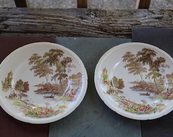 May include: Two antique, off-white ceramic plates with a brown landscape scene featuring trees, a river, and buildings. The plates have a scalloped edge and are displayed on a wooden surface.