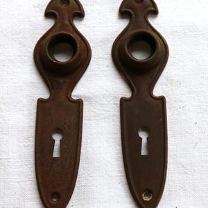 May include: Two antique, ornate, brown metal door escutcheons. Each has a keyhole, a circular opening, and a decorative top. The escutcheons are made of a dark, aged metal, showing signs of wear.