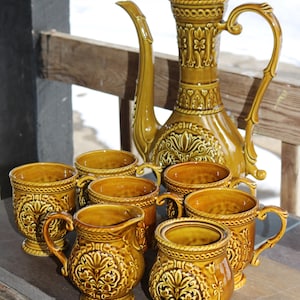 May include: A vintage ceramic coffee set in a warm, golden-brown hue. The set includes a tall, ornate coffee pot with a lid, and several matching mugs and a creamer. The pieces feature detailed floral and scrollwork designs.