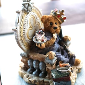 Puede incluir: Figurita detallada de un oso de peluche rey sentado en un trono decorativo. El oso lleva una corona y sostiene un plato pequeño con una porción de pastel. El trono es ornamentado, con una pila de libros en la base. La figurita tiene una estética vintage.