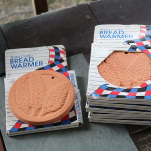May include: A round terracotta bread warmer with a wheat stalk design and a small hole near the edge. The item is packaged in a white box with blue text reading Terra Cotta BREAD WARMER and a red, white, blue, and black checkered pattern.