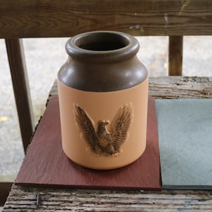Op de afbeelding: Een beige keramische pot met een bruine rand en een gedetailleerd adelaarsreliëf. De pot staat op een donkerrode vierkante tegel. Het is een decoratief item.