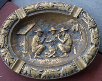 Cenicero de latón macizo de los años 40 con escena de fogata en un pueblo y guitarra fumando.