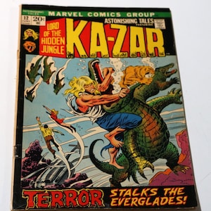 Può includere: Una copertina di fumetto che mostra un uomo con i capelli biondi che combatte un grande alligatore in una palude. La copertina è intitolata "Astonishing Tales Featuring Ka-Zar, Lord of the Hidden Jungle" e il motto è "Terror Stalks the Everglades!"