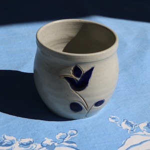 May include: A white ceramic mug with a blue and white tulip design. The mug is sitting on a blue and white tablecloth.