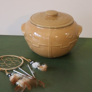 Peut inclure: Un pot à biscuits en céramique beige avec un couvercle et deux poignées. Le pot a un motif géométrique sur les côtés. Un attrape-rêves avec des plumes brunes est au premier plan.