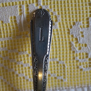 May include: Close-up of a silver spoon with the letter "L" engraved on the handle. The spoon has ornate detailing along the edges and a decorative design at the top. The background is a yellow and white patterned tablecloth.