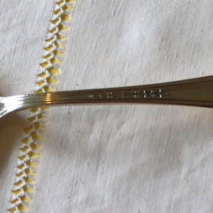 May include: A silver spoon with a reflective surface, resting on a white cloth. The spoon's handle has a decorative floral design at the end and text near the bowl. The spoon is a classic utensil.