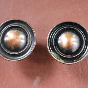 Op de afbeelding: Twee antieke bronzen kastknoppen. Elke knop heeft een centrale, koperkleurige bol omgeven door concentrische zwarte ringen. De knoppen staan tegen een bruine achtergrond, wat hun ontwerp en afwerking benadrukt.