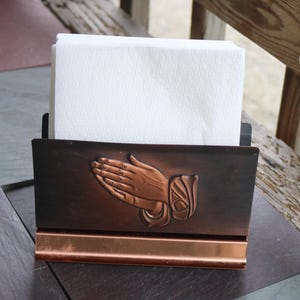 May include: A copper-toned metal napkin holder with a decorative relief of praying hands. The holder is rectangular with a copper-colored base and holds a stack of white paper napkins. The design is vintage and suitable for a dining table.