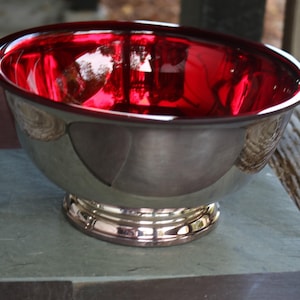 Może przedstawiać: Dekoracyjna srebrna misa z żywym czerwonym wnętrzem. Misa ma szeroki, zaokrąglony kształt i stoi na okrągłej, złotej podstawie. Odbijająca światło powierzchnia misy tworzy uderzający efekt wizualny.