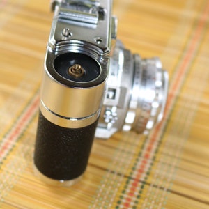 Pu&ograve; includere: Una macchina fotografica vintage da 35 mm in argento e nero con un copriobiettivo sul davanti. La macchina fotografica &egrave; appoggiata su un tappetino di bamb&ugrave; intrecciato.