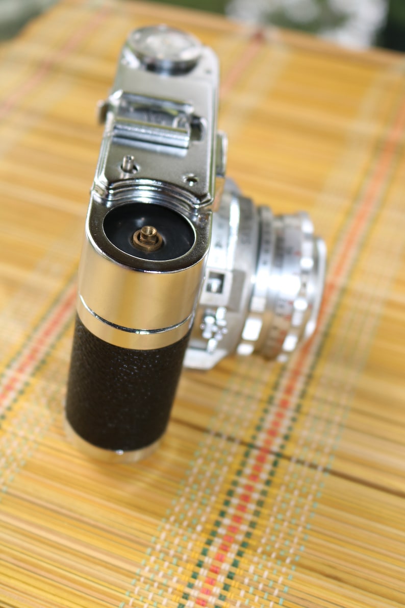 Pu&ograve; includere: Una macchina fotografica vintage da 35 mm in argento e nero con un copriobiettivo sul davanti. La macchina fotografica &egrave; appoggiata su un tappetino di bamb&ugrave; intrecciato.