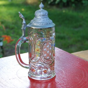 May include: A clear glass beer stein with a pewter lid and colourful floral designs. The stein has a handle and a round body. The lid has a decorative design.