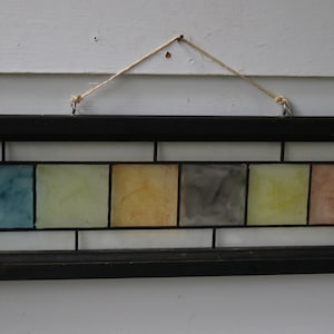 May include: A rectangular stained glass panel framed in dark wood, featuring six square glass panes in shades of blue, light green, yellow, gray, lime green, and peach. The panel is suspended by a thin twine string attached to two metal eyelets.