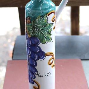 Op de afbeelding: Witte keramische olijfoliefles met een turquoise handvat en tuit. De fles is versierd met een tros paarse druiven, groene bladeren en het woord "Tadinate". Een kurk sluit de opening af.