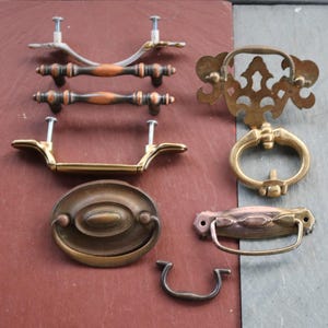 May include: A collection of vintage cabinet hardware, including handles and pulls. The hardware features various designs, including ornate brass, curved metal, and oval shapes. The colors range from gold to bronze and dark brown, with visible screws for mounting.