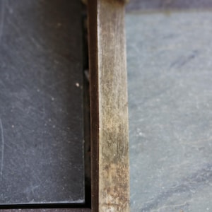 Puede incluir: Primer plano de un formón para carpintería. El formón tiene un mango de madera con una férula de metal y una hoja rectangular biselada. La hoja muestra signos de desgaste. El fondo es una superficie gris oscuro.