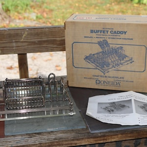 Peut inclure: Un présentoir à buffet argenté avec une base rectangulaire et plusieurs compartiments, à côté de sa boîte en carton d'origine. La boîte affiche le texte "Buffet Caddy" et un schéma de couverts. Une notice imprimée est également visible.