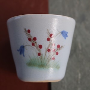 Puede incluir: Una pequeña taza de cerámica blanquecina con un diseño floral pintado a mano. El diseño presenta bayas rojas, campanillas azules y hierba verde. La taza tiene una superficie ligeramente texturizada y una forma sencilla.