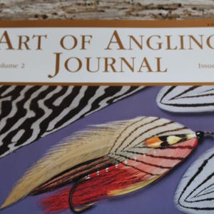 May include: The cover of the "Art of Angling Journal" features a colorful fly fishing lure on a purple background. The journal's title is in gold lettering, with the volume and issue numbers visible.