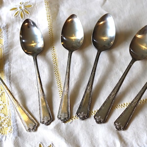 May include: Six vintage silver spoons with oval bowls and ornate handles are arranged on a white and yellow patterned tablecloth. The spoons are reflective, showing the surrounding environment.