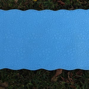 May include: A rectangular, light blue mat with a scalloped edge. The mat has a floral pattern embossed across its surface. The mat is lying on a bed of green grass and fallen leaves.