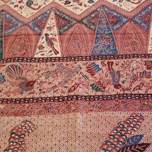 May include: A textile fabric featuring a complex batik pattern in shades of red, blue, and cream. The design includes triangular motifs with floral elements, birds, and intricate geometric borders. The surface is covered in detailed, repeating patterns.