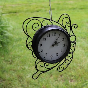 May include: A black metal outdoor clock with a white face and black numerals. The clock is suspended by a metal hook and is surrounded by a decorative, butterfly-shaped frame. The background is blurred green grass.