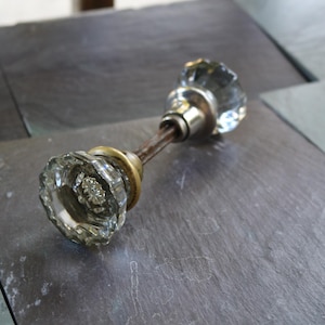 May include: A clear, faceted glass door knob set featuring a central metal spindle. The two glass knobs are connected by a thin, weathered metal rod, with one knob attached to a brass-toned circular base and the other to a silver-toned base.