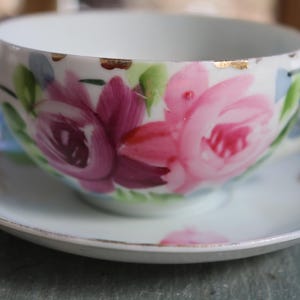 May include: White teacup and saucer set with hand-painted pink and purple roses and green leaves. The cup has a gold rim and a curved handle. The saucer has a matching floral design and gold trim. A vintage tea set.