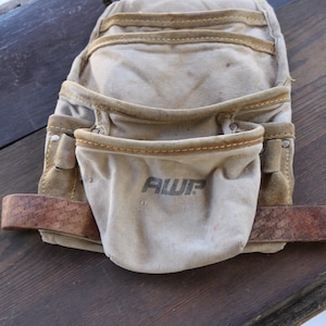 May include: A tan canvas tool belt with multiple pockets and a brown leather belt. The front pocket has the letters "AWP" printed on it. The tool belt is designed for carrying tools.