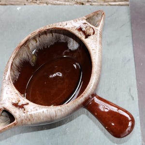 May include: A brown and white glazed ceramic gravy boat with a handle. The boat has a brown interior and a white exterior with brown speckles.