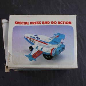 May include: A toy airplane in a box. The plane is white and blue with red stripes and the text "HW-303". The box reads "SPECIAL PRESS AND GO ACTION". The plane has black wheels and blue accents.