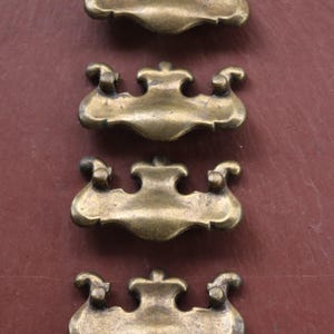 May include: Four antique brass drawer pulls with a decorative design. Each pull has a curved shape with ornate detailing, including scrolled ends and a central flourish. The pulls are arranged in a vertical row against a brown background.