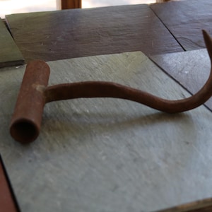 May include: A rustic, weathered iron hay hook featuring a cylindrical T-shaped handle and a curved, pointed metal prong. The tool displays a uniform, oxidized brown patina across its entire surface, indicating age and heavy-duty use.