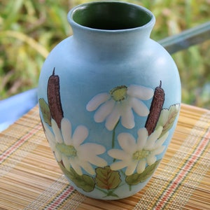 Puede incluir: Un jarrón de cerámica azul claro con un borde verde, decorado con flores blancas y tallos marrones. El jarrón está sobre una estera de bambú tejida.