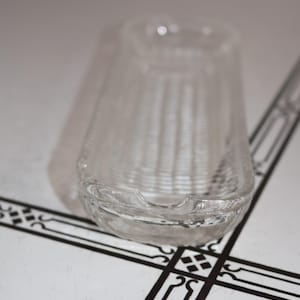 May include: A clear glass ashtray with a rectangular shape and a textured, ribbed design. The ashtray has a small, curved indentation on one side for resting cigarettes. The glass is transparent, allowing light to pass through.