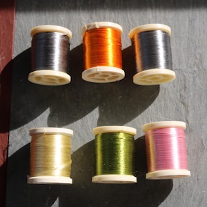 May include: Six spools of metallic thread in various colours, including silver, orange, and pink. The spools are arranged on a grey surface, with a contrasting red-brown area in the background. The thread is wound on white plastic spools.