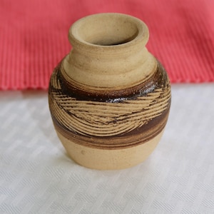 May include: A small, brown and tan ceramic vase with a decorative, striped pattern. The vase is made of earthenware and has a rounded shape.