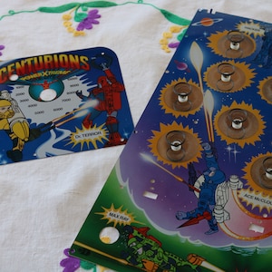 May include: Two game boards for a pinball machine featuring the Centurions characters. The top board is blue with a yellow and white scorecard and features the characters Jake Rockwell and Dr. Terror. The bottom board is blue with a yellow, orange, and white design and features the characters Max Ray, Ace McCloud, and Strafer.