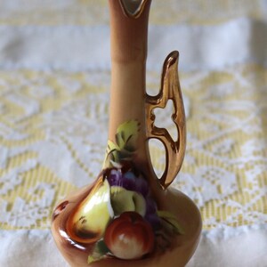 May include: A small, tan-colored ceramic vase with a gold handle and rim. The vase is decorated with a colorful still life of fruits, including apples, pears, and grapes. The vase has a unique shape and is likely used for decorative purposes.